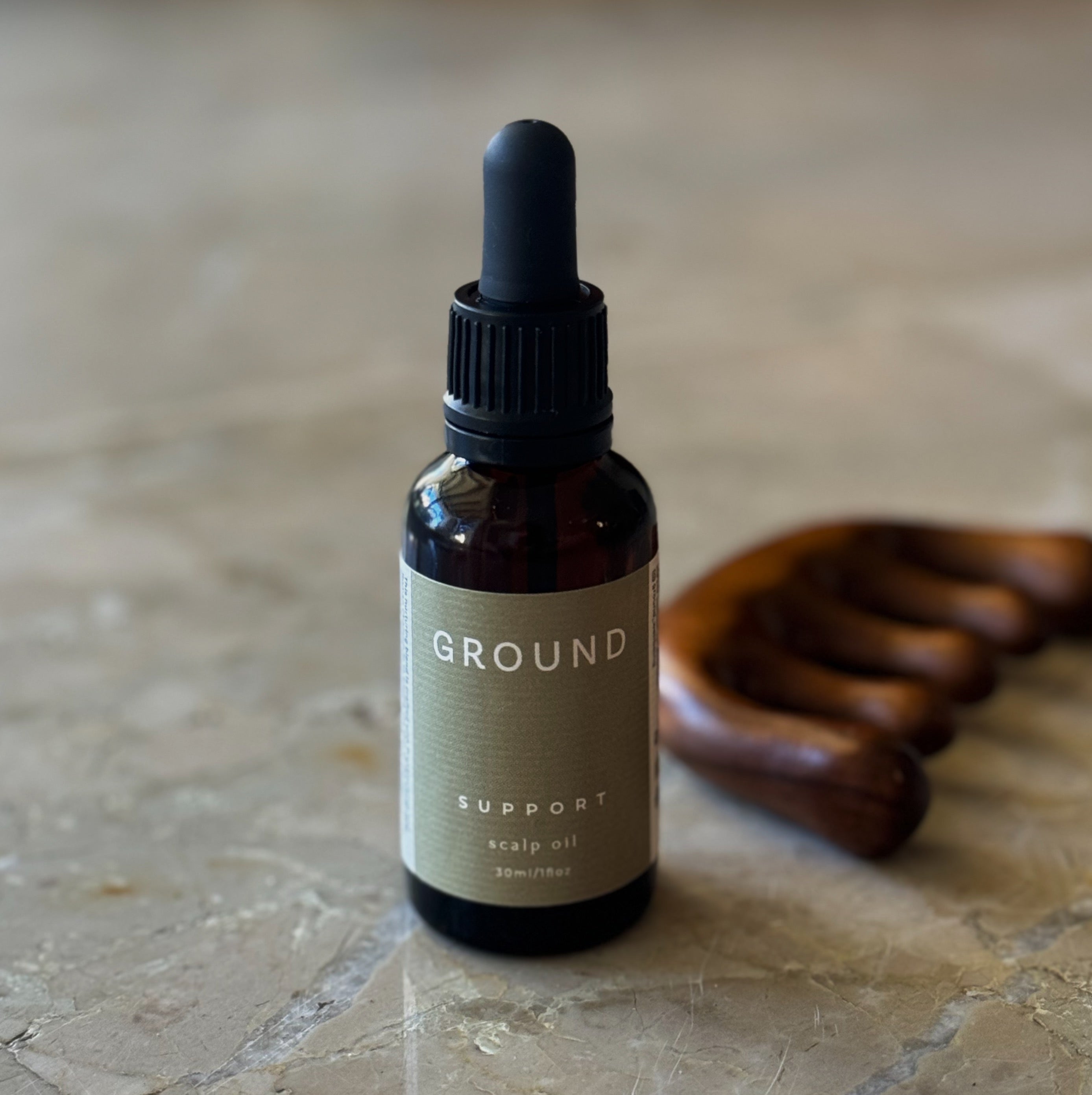 Photograph of a glass bottle of Ground Support Scalp Oil 30ml sitting beside a wooden wide tooth comb on a marble countertop.