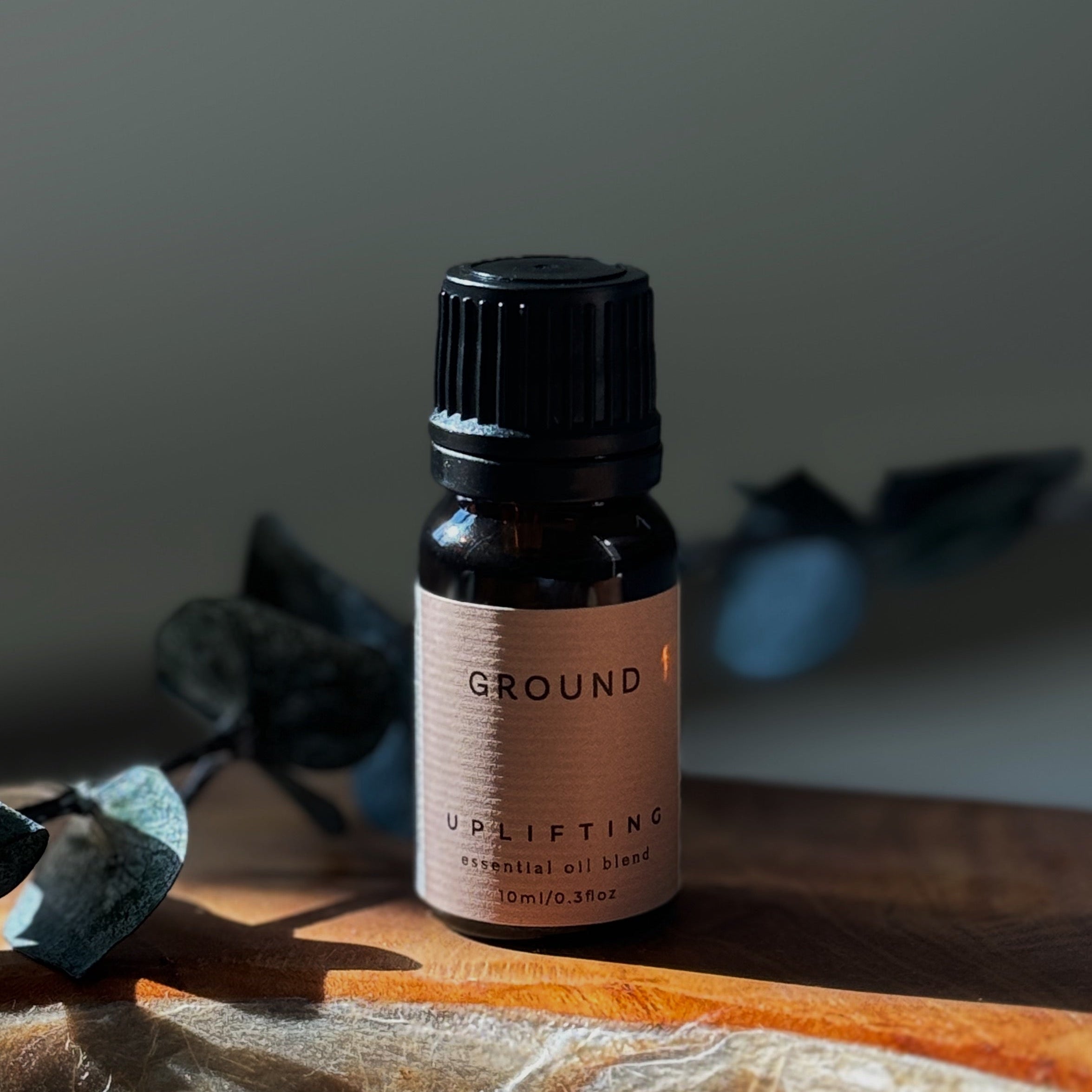 Bottle of 'GROUND' uplifting essential oil blend on a wooden surface with blurred eucalyptus leaves in the background.
The bottle is a small brown glass bottle with a cream label stating 'GROUND' uplifting essential oil blend 10ml, 0.3 floz.