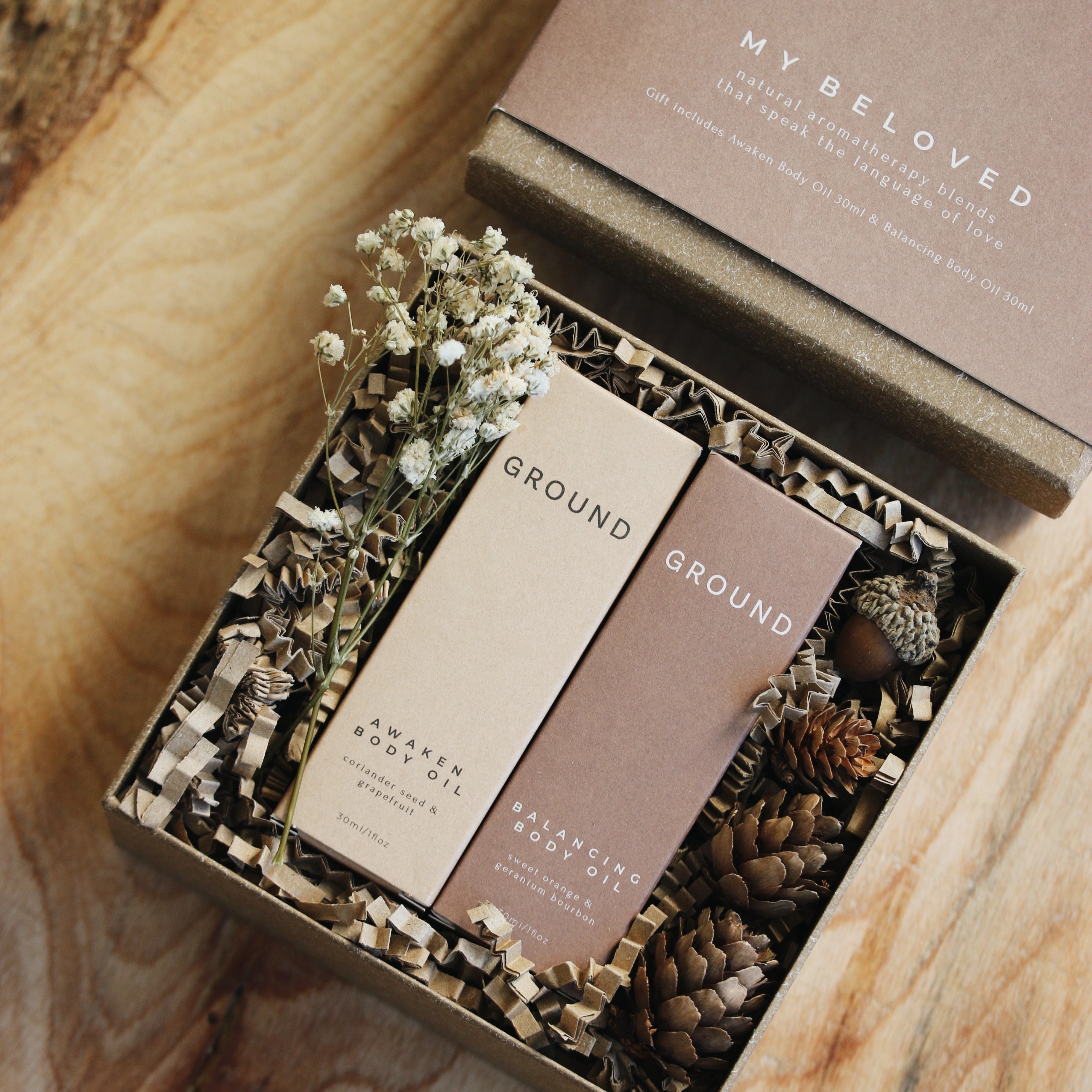 Image of Ground My Beloved gift set contents. Photograph shows premium cardboard packaging filled with sustainable cardboard stuffing surrounding two bottles of body oil in their own cardboard presentation boxes. The Awaken Body Oil contains coriander seed and grapefruit, whilst the balancing oil contains sweet orange and geranium bourbon. The contents are also decorated with small white natural dried flowers.