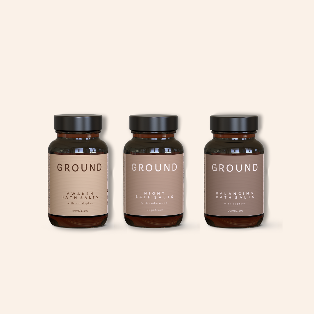 Three bottles of 'GROUND' bath salts on a beige background. The image shows Ground Wellbeing The Bathing Rituals Trio contents. It contains Awaken Bath Salts with eucalyptus, Night Bath Salts with cedarwood and Balancing Bath Salts with cypress.