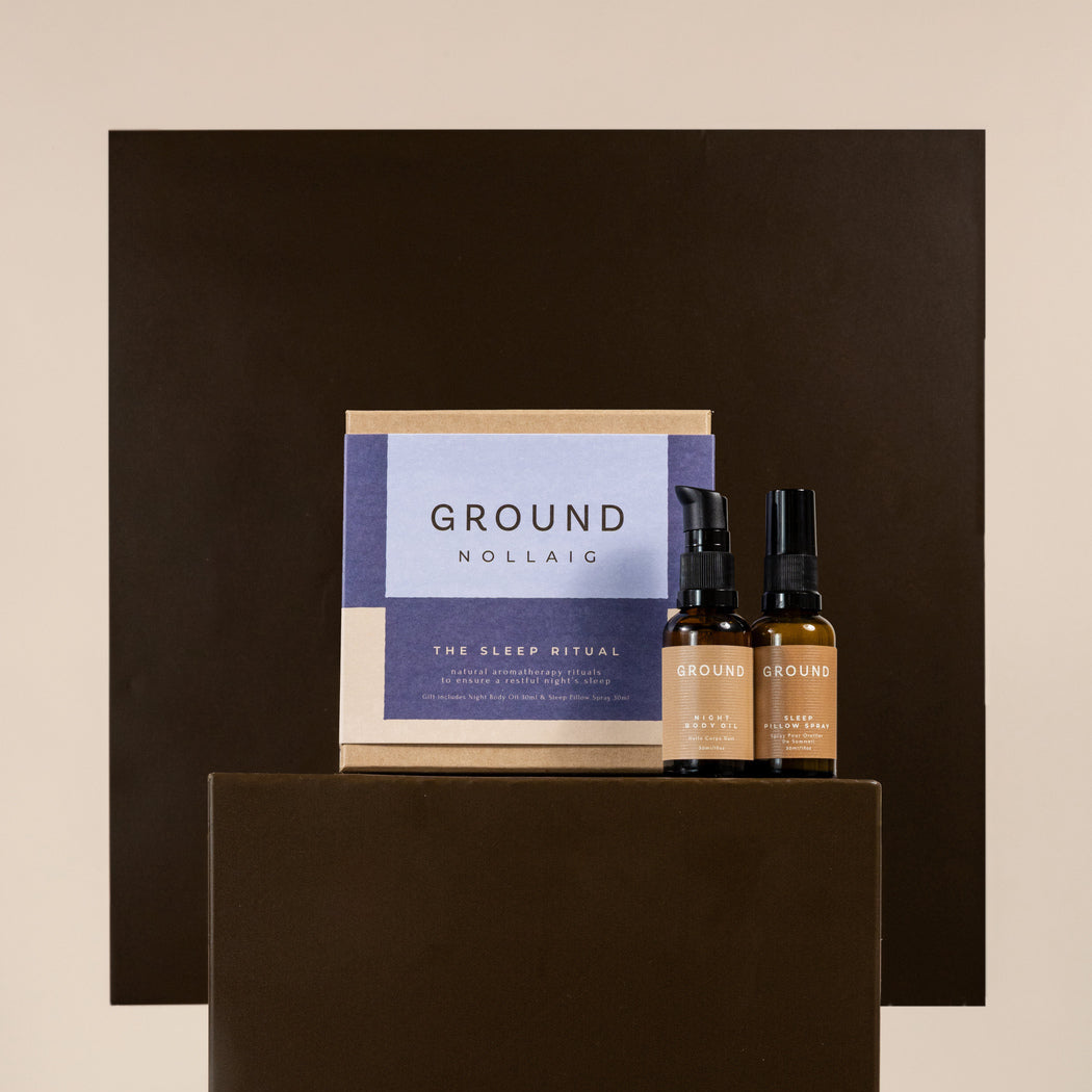 A boxed set and two small amber bottles with black droppers displayed on a dark brown pedestal against a beige background. The front label reads GROUND NOLLAG sleep ritual