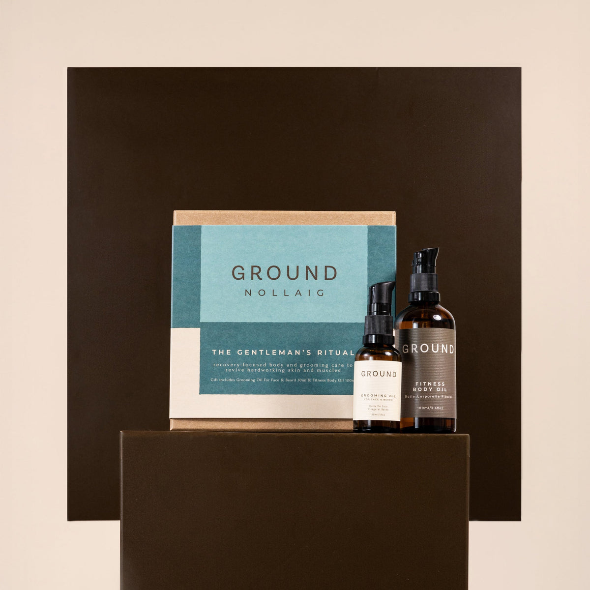 A boxed grooming set from Ground Nollaig displayed on a dark pedestal against a warm beige background. The set includes a teal and cream box labeled 'GROUND NOLLAIG' and two dark amber bottles with black caps and white labels.