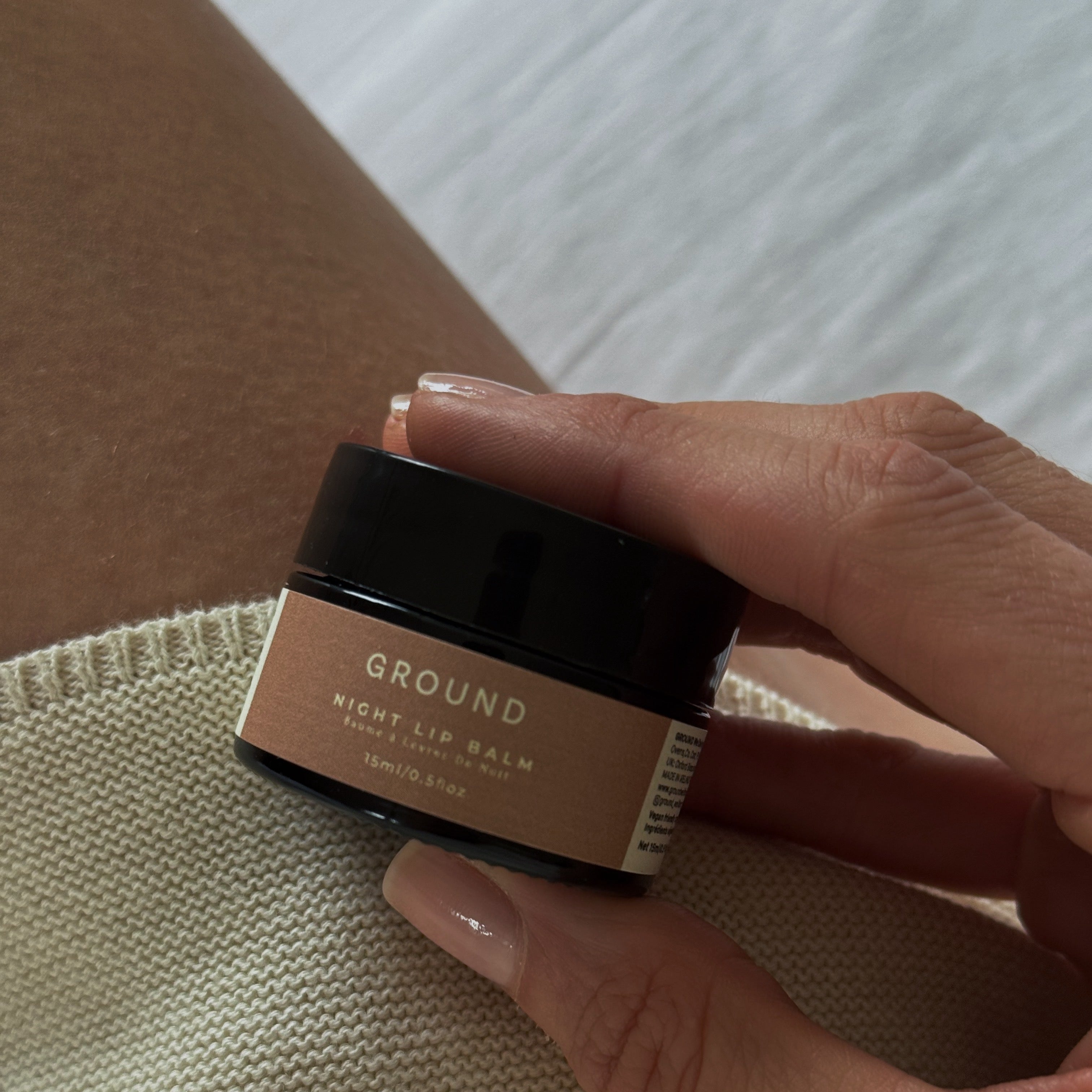 A hand holding a small jar of GROUND Night Lip Balm over a textured beige knit fabric, with a neutral background.