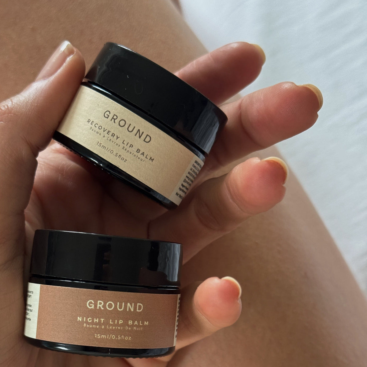 A hand holds two small cosmetic jars labeled GROUND Recovery Lip Balm and GROUND Night Lip Balm against a softly blurred skin and light fabric background.