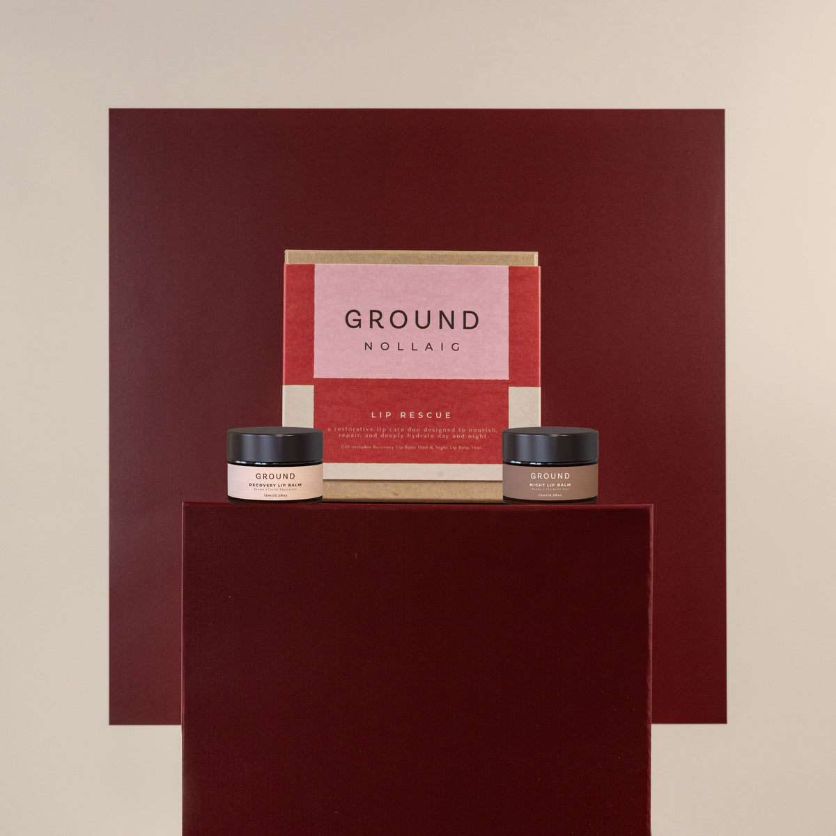A cosmetic gift set from Ground on a dark red pedestal against a beige background. The set includes a pink and red box labeled 'GROUND LIP RESCUE' and two small jars with black lids.