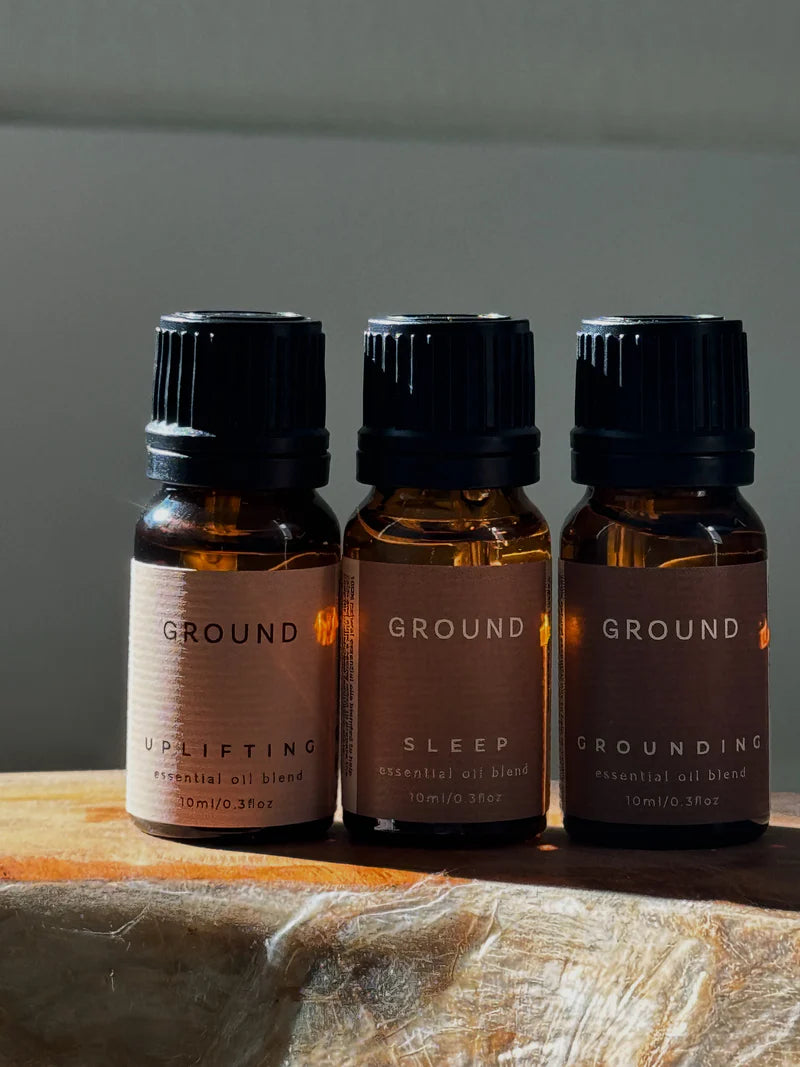 Three essential oil bottles labelled 'Ground', 'Uplifting', 'Sleep', and 'Grounding' on a wooden surface with a neutral background.