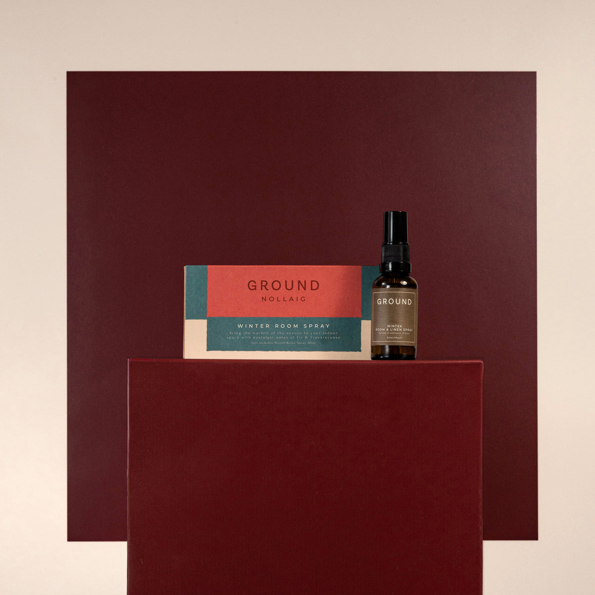 A bottle of Ground Winter Room Spray and its box displayed on a dark red pedestal against a matching dark red background panel and a light beige wall.